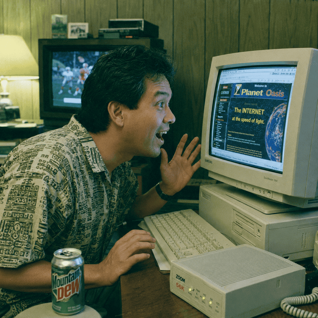 A man in a patterned short-sleeve shirt leans toward a beige 1990s CRT monitor with his mouth open in surprise. The screen shows a colorful web page reading 'Welcome to Planet Oasis: The INTERNET at the speed of light.' On the desk sit a beige tower PC, a Zoom 56K external modem with blinking green lights, and a can of Mountain Dew. Wood paneling and a CRT television playing a football game fill the background.